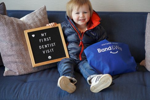 A toddler sitting in a big chair at a dental office, holding a sign that says, "My first dentist visit." He is smiling big and holds a Band & Wire merchandise bag.